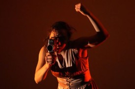 A dancer pointng a camcorder towards the viewer with her other arm raised in a loose fist.